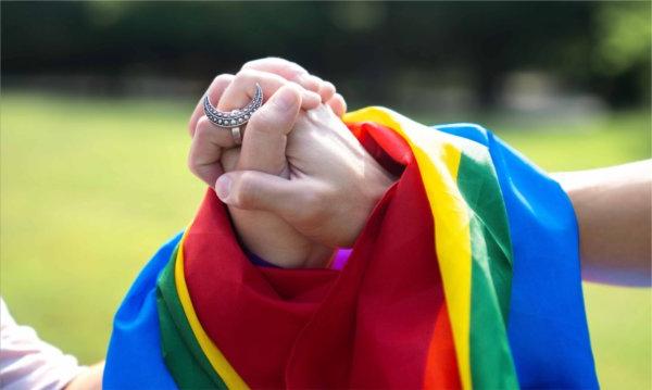  Two hands clasped together are draped in a pride flag.