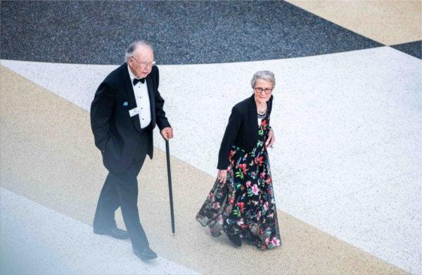  A couple wearing a tuxedo and floral gown are seen from a balcony walking in to an event. 